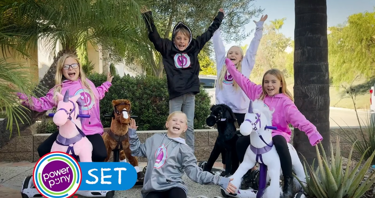 Group of excited kids in Power Pony hoodies posing with Power Pony toys outdoors.