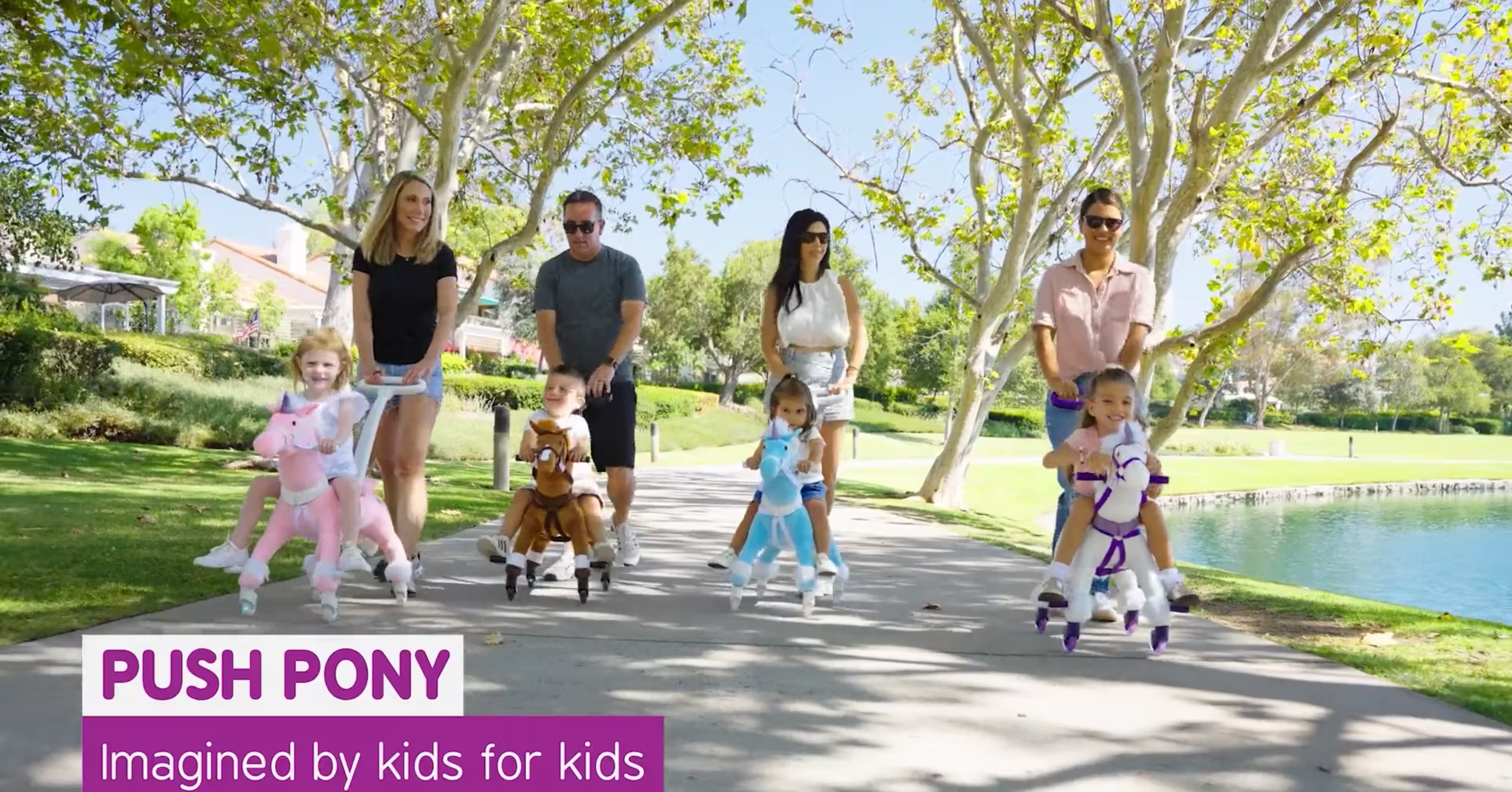 A group of children riding different colored Power Ponies, each being pushed by adults on a sunny park path. Text reads "Push Pony: Imagined by kids for kids.