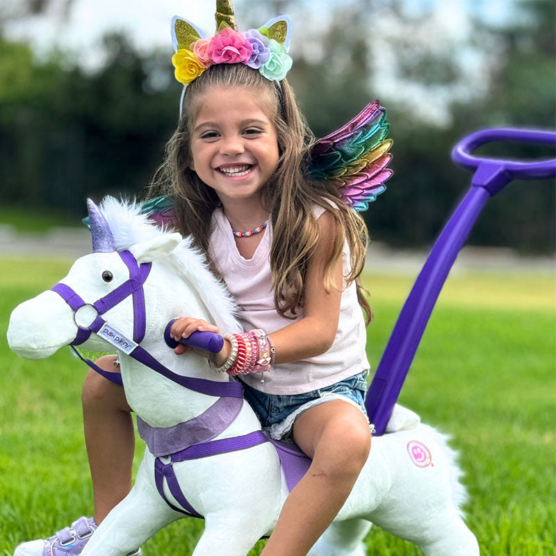 Smiling girl in unicorn headband and wings riding a white Push Pony with a handlebar.