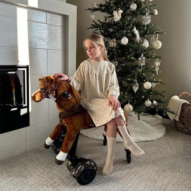 Young girl sitting on a brown Power Pony next to a decorated Christmas tree indoors.