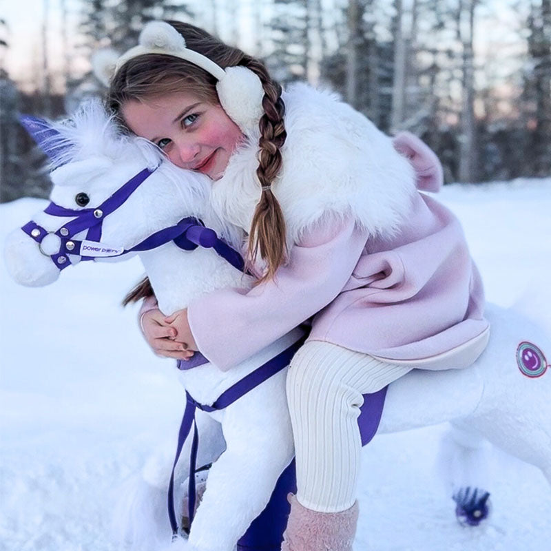 Girl in winter clothing hugging a white Power Pony unicorn in a snowy setting.