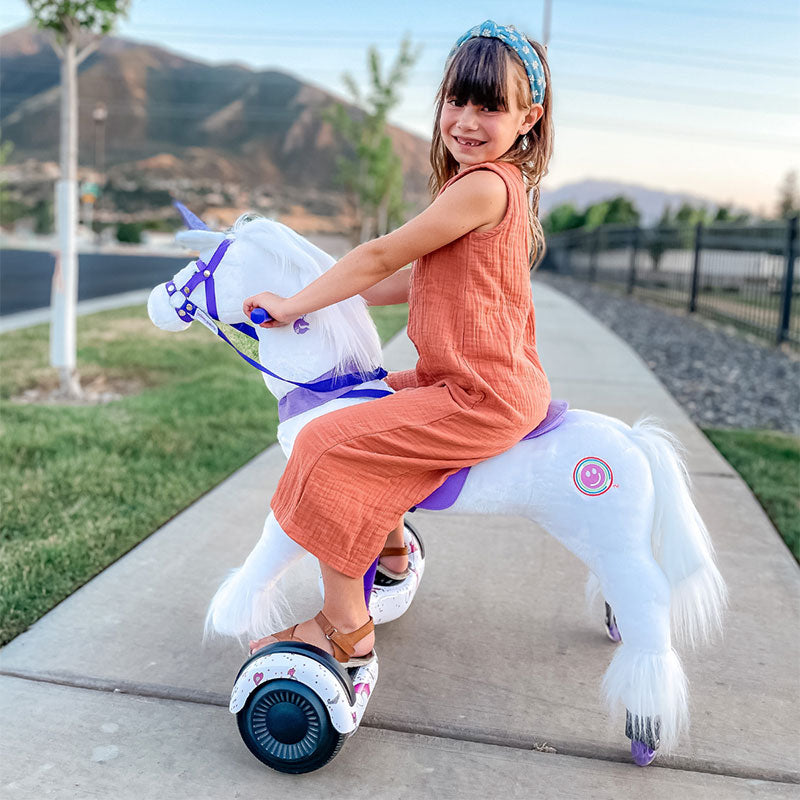 Smiling girl in an orange dress riding a white Power Pony unicorn on a sidewalk.