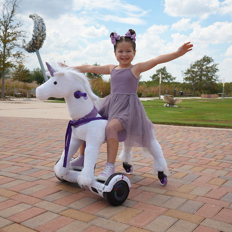 Young girl in a purple dress riding a white Power Pony unicorn with arms outstretched on a brick pathway.