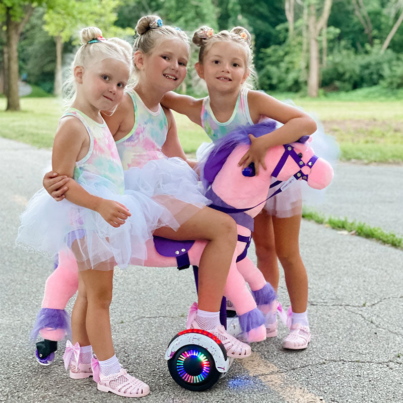 Three young girls in tutus pose with a pink Power Pony outdoors.